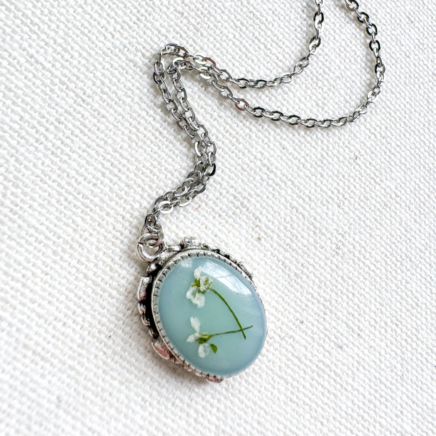 Handcrafted pressed flower pendant necklace featuring real dried Queen Anne's lace flowers against aquamarine backdrop, set in an ornate vintage silver frame. Perfect for nature-inspired jewelry lovers.