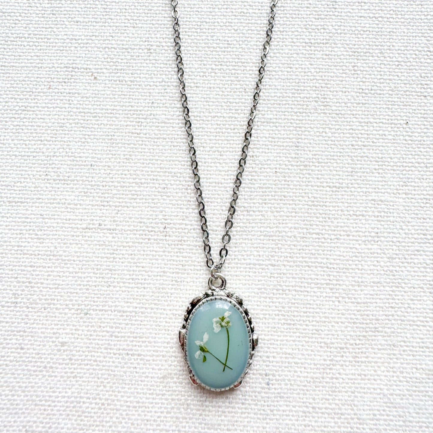 Handcrafted pressed flower pendant necklace featuring real dried Queen Anne's lace flowers against aquamarine backdrop, set in an ornate vintage silver frame. Perfect for nature-inspired jewelry lovers.