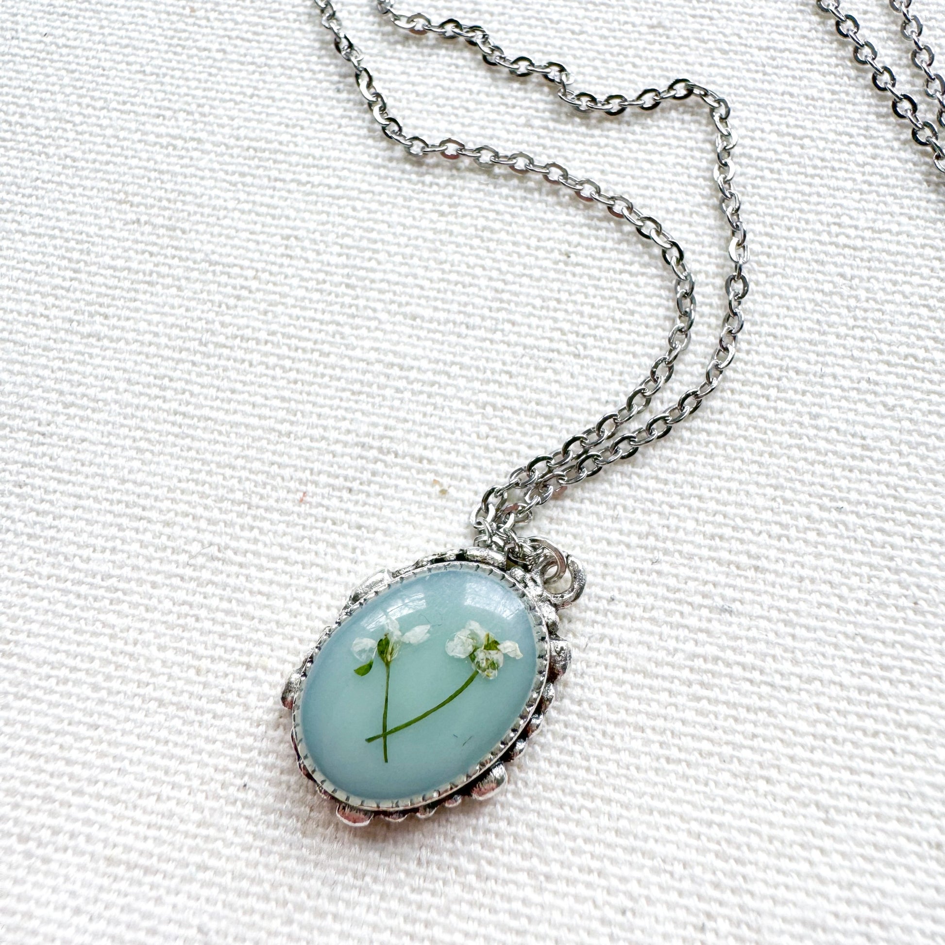 Handcrafted pressed flower pendant necklace featuring real dried Queen Anne's lace flowers against aquamarine backdrop, set in an ornate vintage silver frame. Perfect for nature-inspired jewelry lovers.