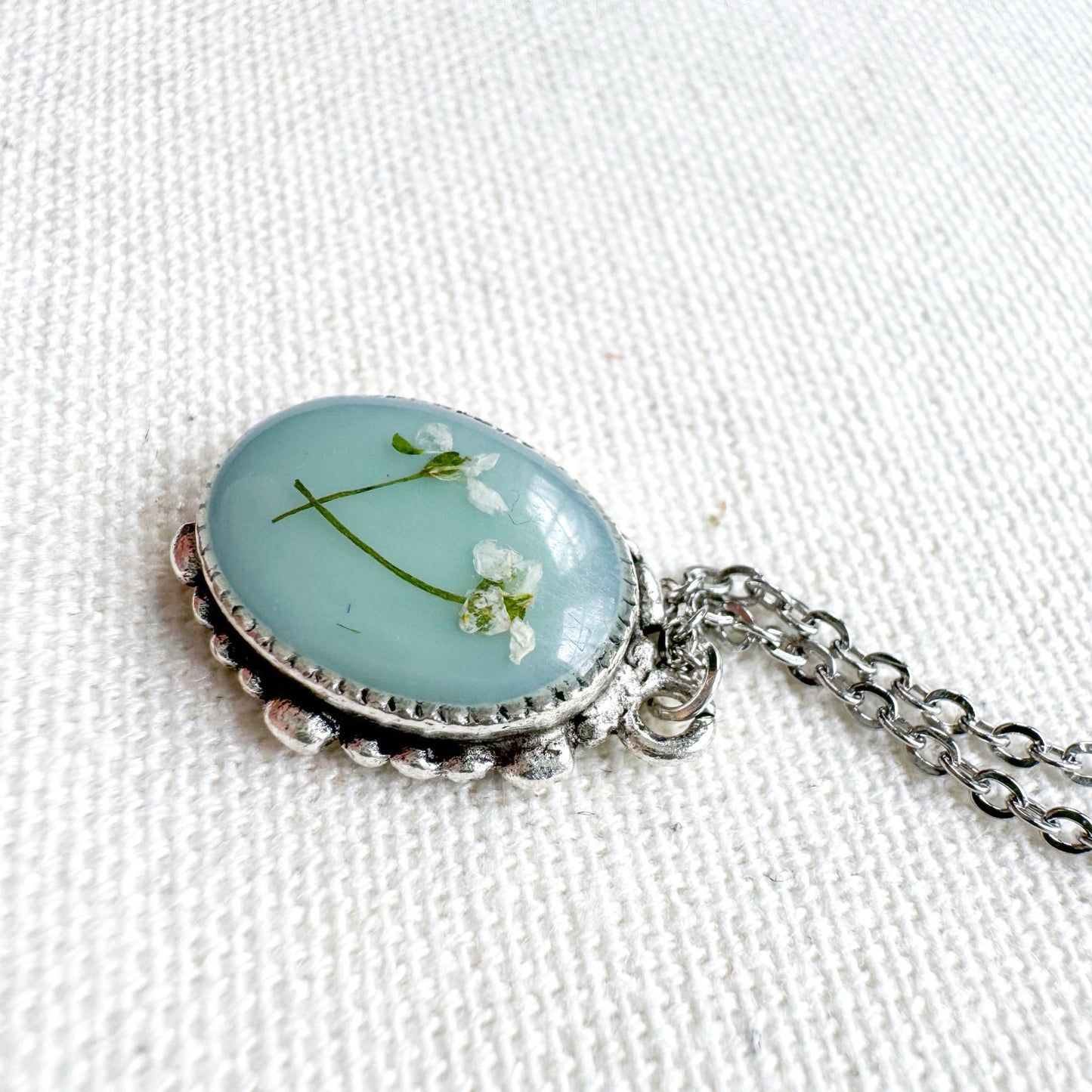 Handcrafted pressed flower pendant necklace featuring real dried Queen Anne's lace flowers against aquamarine backdrop, set in an ornate vintage silver frame. Perfect for nature-inspired jewelry lovers.