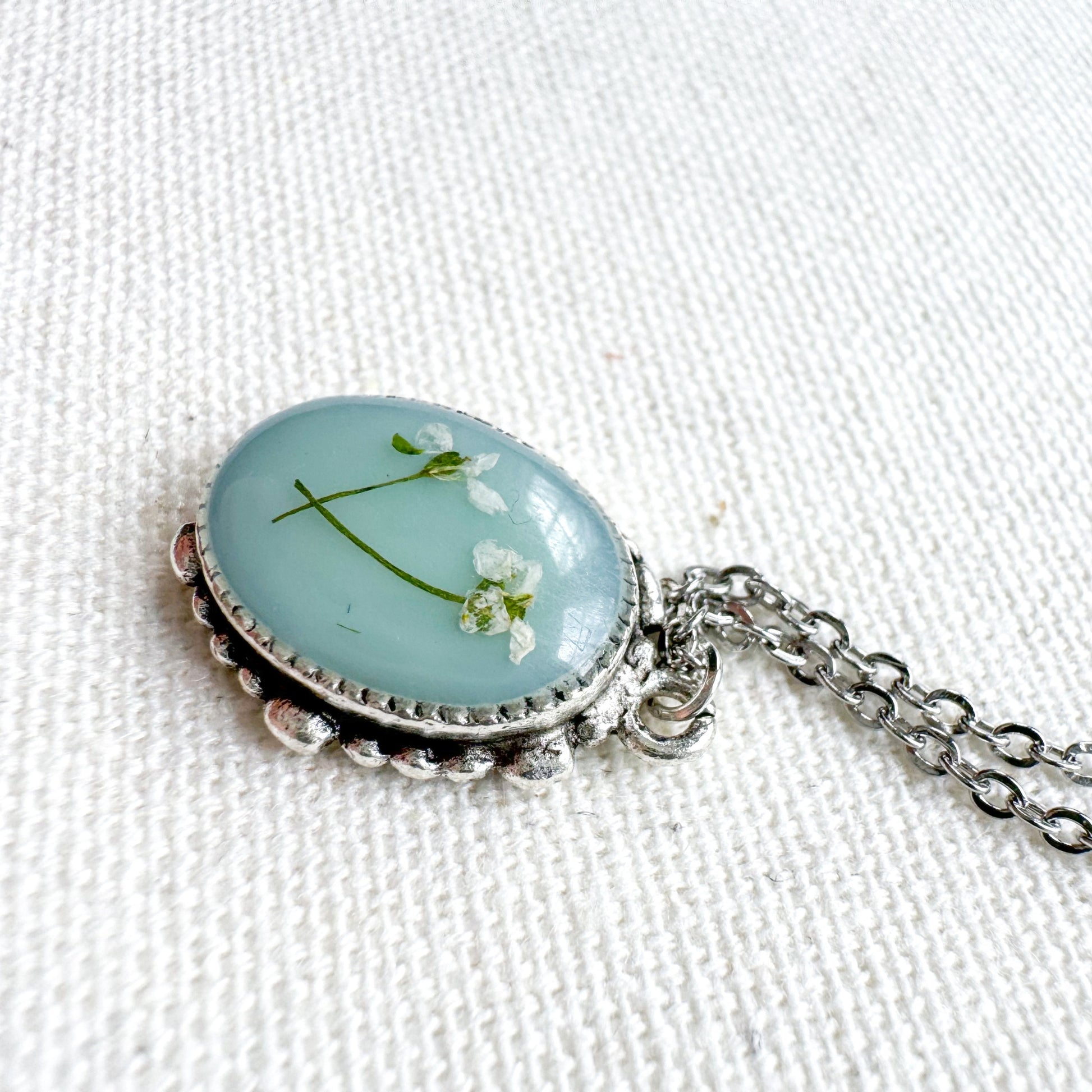 Handcrafted pressed flower pendant necklace featuring real dried Queen Anne's lace flowers against aquamarine backdrop, set in an ornate vintage silver frame. Perfect for nature-inspired jewelry lovers.