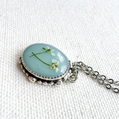 Handcrafted pressed flower pendant necklace featuring real dried Queen Anne's lace flowers against aquamarine backdrop, set in an ornate vintage silver frame. Perfect for nature-inspired jewelry lovers.