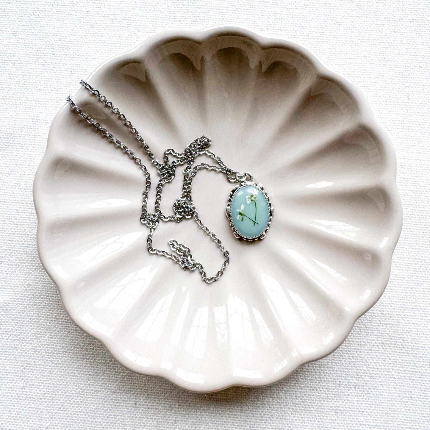 Handcrafted pressed flower pendant necklace featuring real dried Queen Anne's lace flowers against aquamarine backdrop, set in an ornate vintage silver frame draped on a ceramic flower trinket tray.
