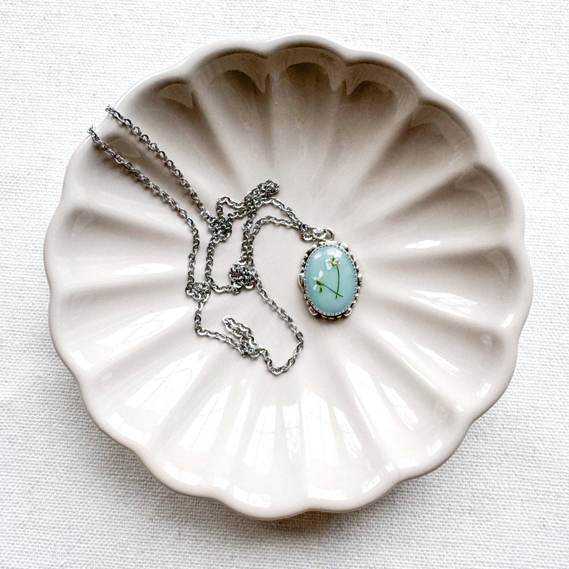 Handcrafted pressed flower pendant necklace featuring real dried Queen Anne's lace flowers against aquamarine backdrop, set in an ornate vintage silver frame draped on a ceramic flower trinket tray.