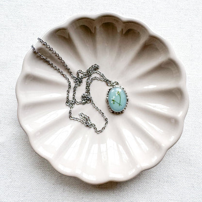 Handcrafted pressed flower pendant necklace featuring real dried Queen Anne's lace flowers against aquamarine backdrop, set in an ornate vintage silver frame draped on a ceramic flower trinket tray.