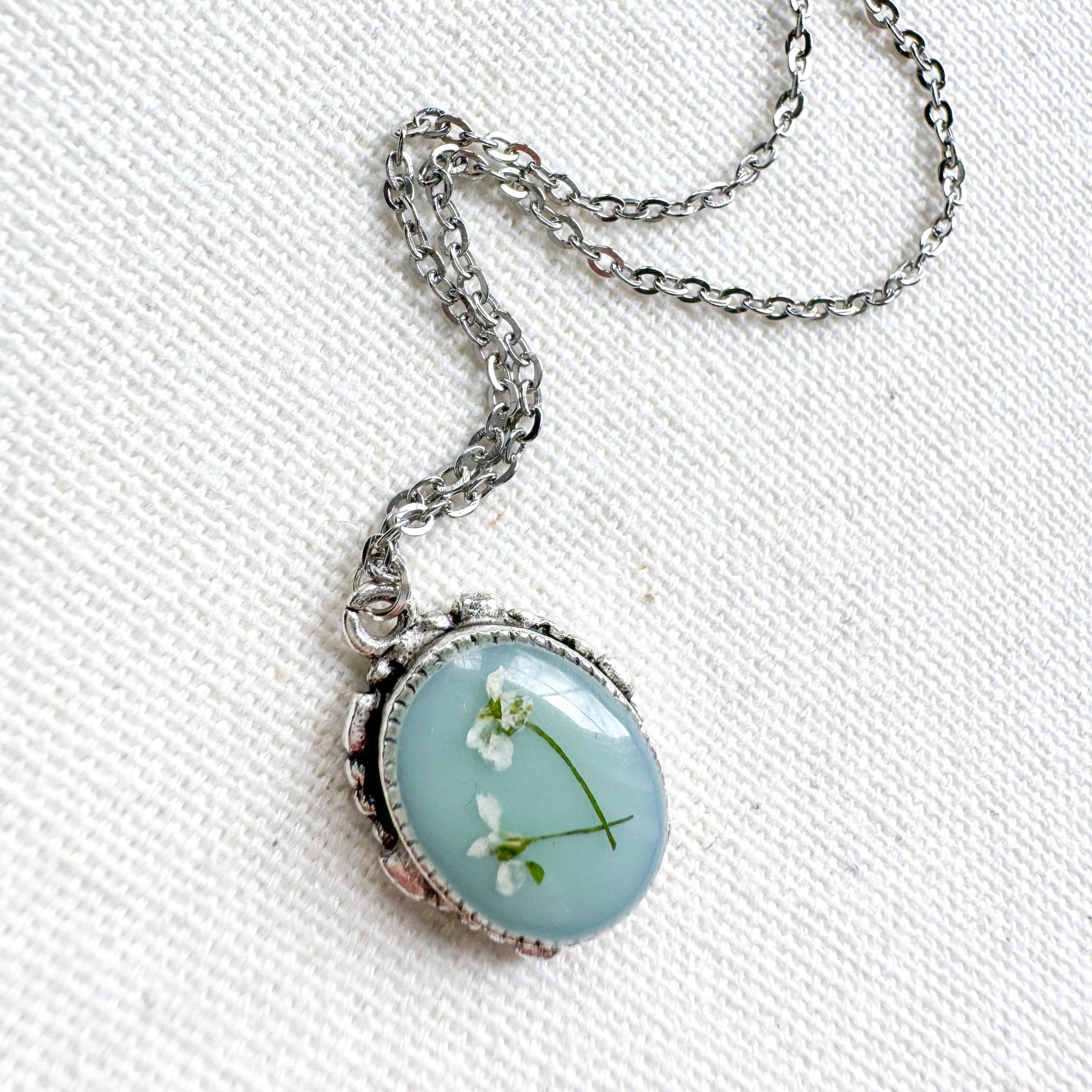 Handcrafted pressed flower pendant necklace featuring real dried Queen Anne's lace flowers against aquamarine backdrop, set in an ornate vintage silver frame. Perfect for nature-inspired jewelry lovers.