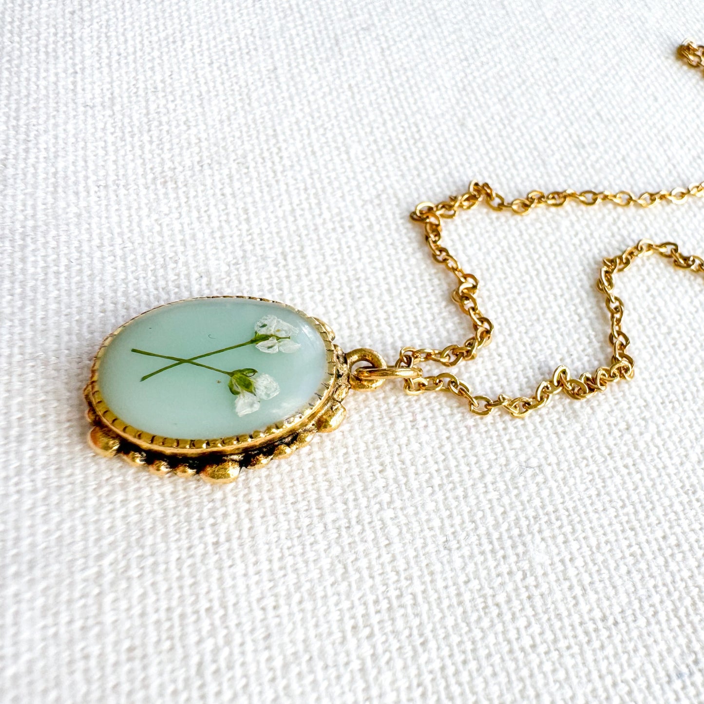 Handcrafted pressed flower pendant necklace featuring real dried Queen Anne's lace flowers against aquamarine backdrop, set in an ornate vintage gold frame, perfect for nature-inspired jewelry lovers.