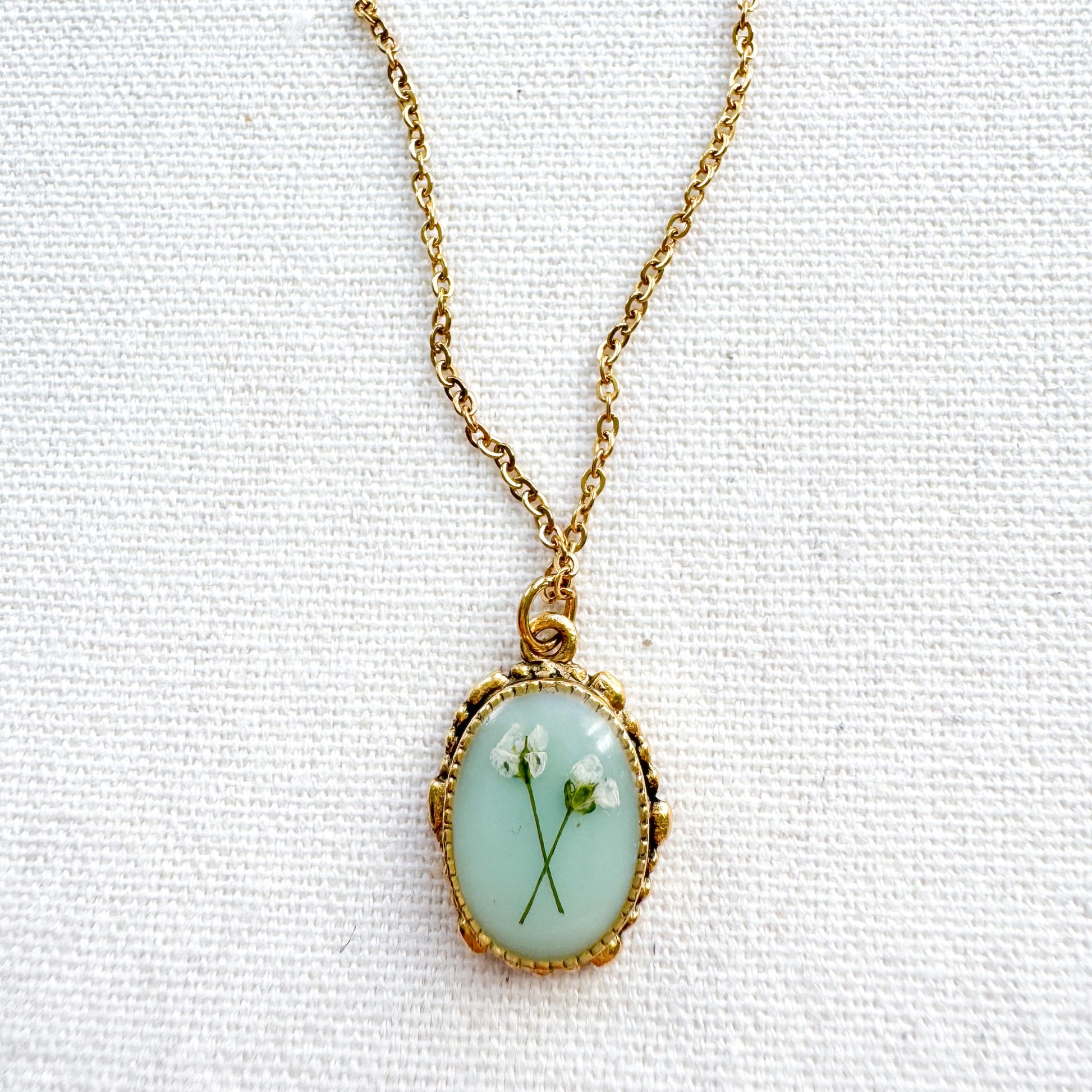 Handcrafted pressed flower pendant necklace featuring real dried Queen Anne's lace flowers against aquamarine backdrop, set in an ornate vintage gold frame, perfect for nature-inspired jewelry lovers.