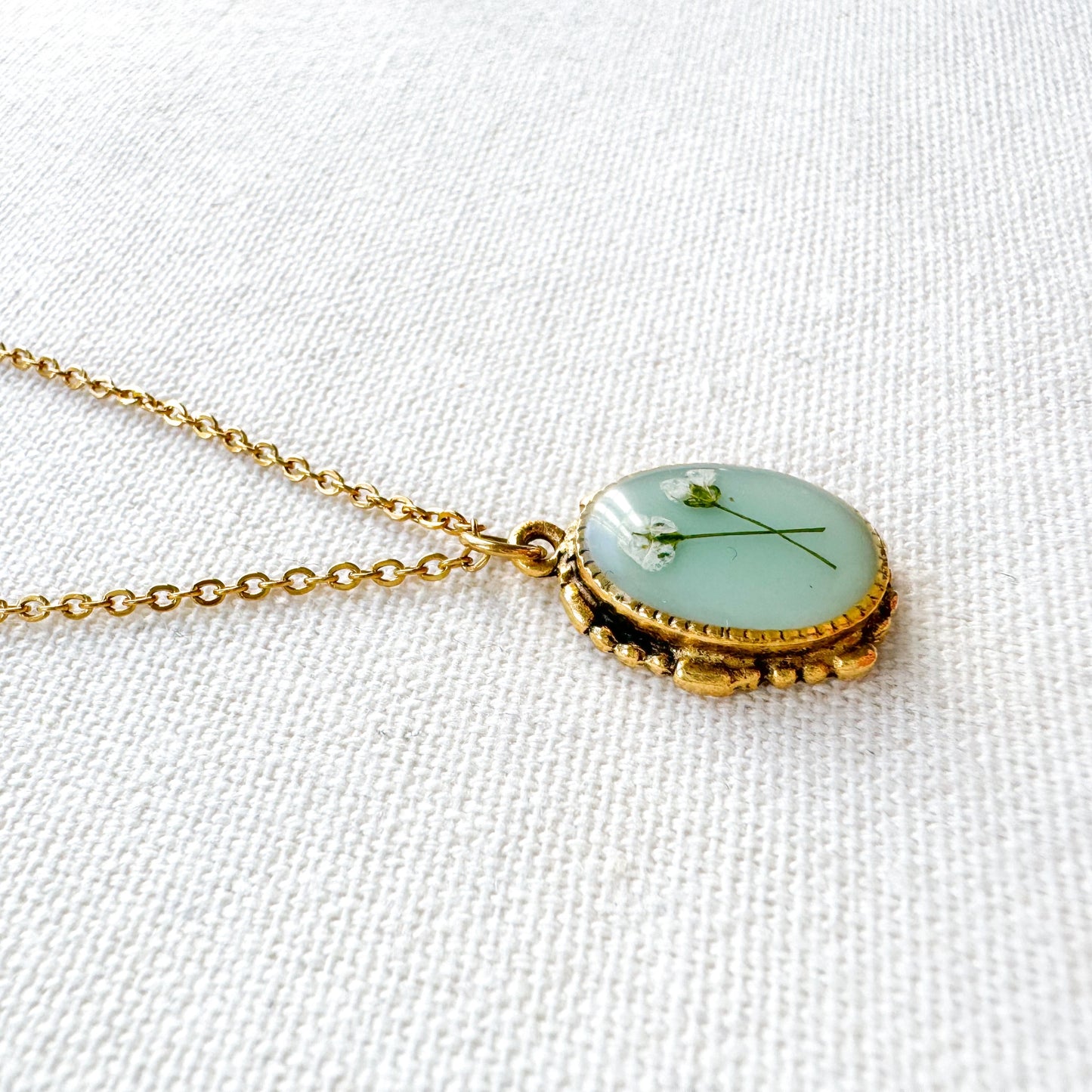Handcrafted pressed flower pendant necklace featuring real dried Queen Anne's lace flowers against aquamarine backdrop, set in an ornate vintage gold frame, perfect for nature-inspired jewelry lovers.