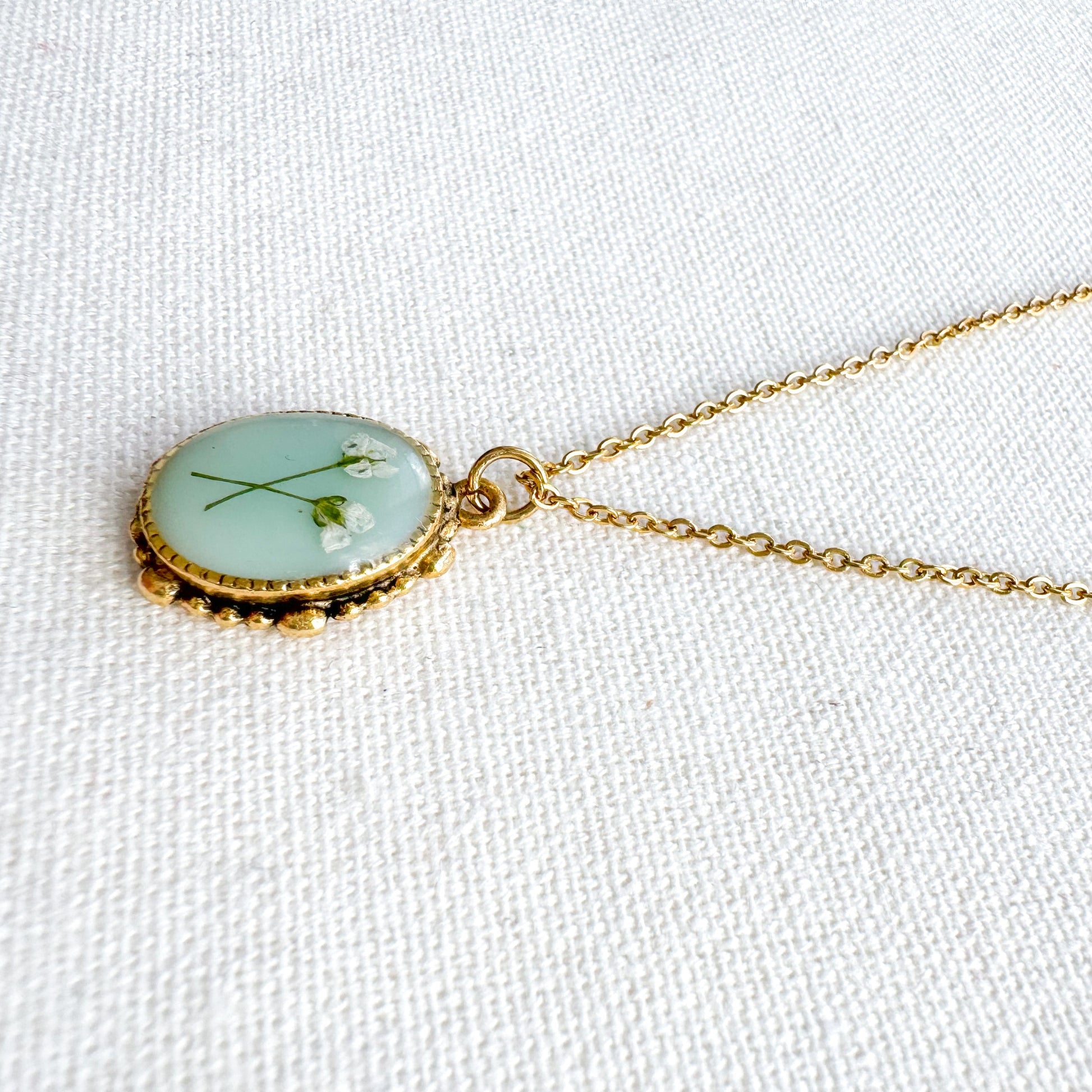 Handcrafted pressed flower pendant necklace featuring real dried Queen Anne's lace flowers against aquamarine backdrop, set in an ornate vintage gold frame, perfect for nature-inspired jewelry lovers.