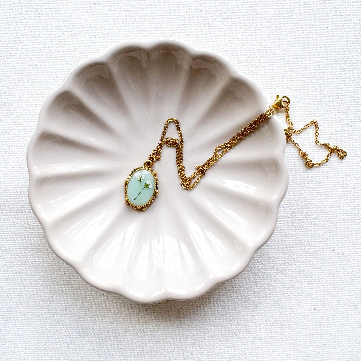 Handcrafted pressed flower pendant necklace featuring real dried Queen Anne's lace flowers against aquamarine backdrop, set in an ornate vintage gold frame, draped across a ceramic trinket tray.