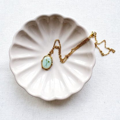 Handcrafted pressed flower pendant necklace featuring real dried Queen Anne's lace flowers against aquamarine backdrop, set in an ornate vintage gold frame, draped across a ceramic trinket tray.