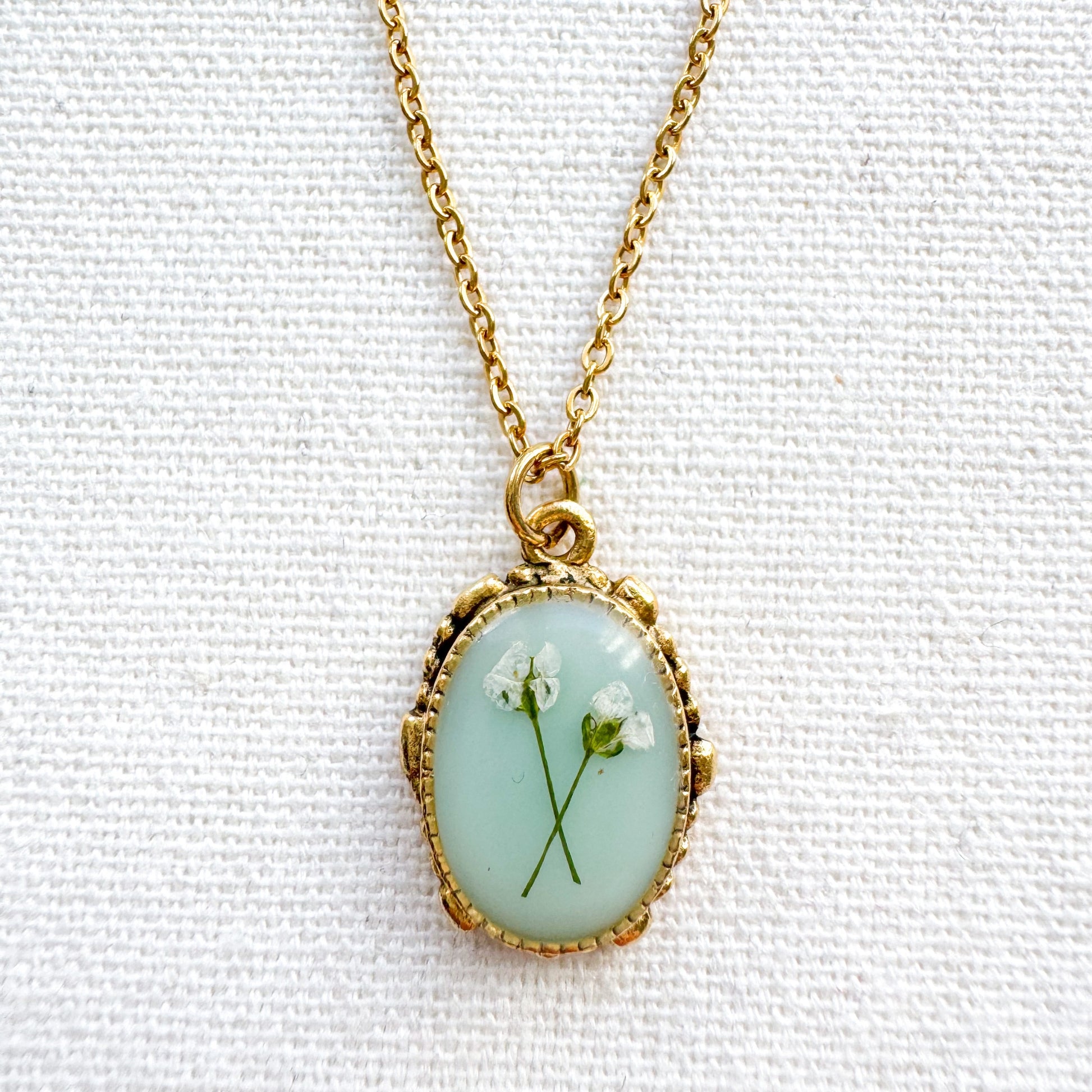 Handcrafted pressed flower pendant necklace featuring real dried Queen Anne's lace flowers against aquamarine backdrop, set in an ornate vintage gold frame, perfect for nature-inspired jewelry lovers.