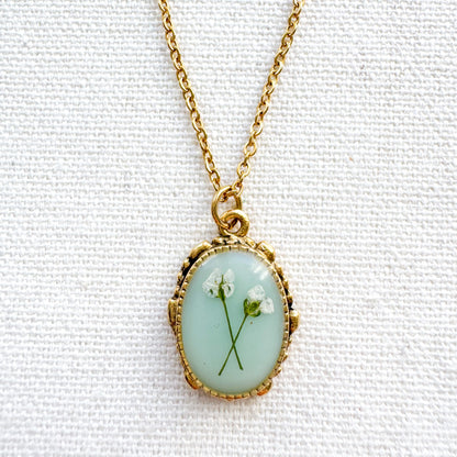 Handcrafted pressed flower pendant necklace featuring real dried Queen Anne's lace flowers against aquamarine backdrop, set in an ornate vintage gold frame, perfect for nature-inspired jewelry lovers.