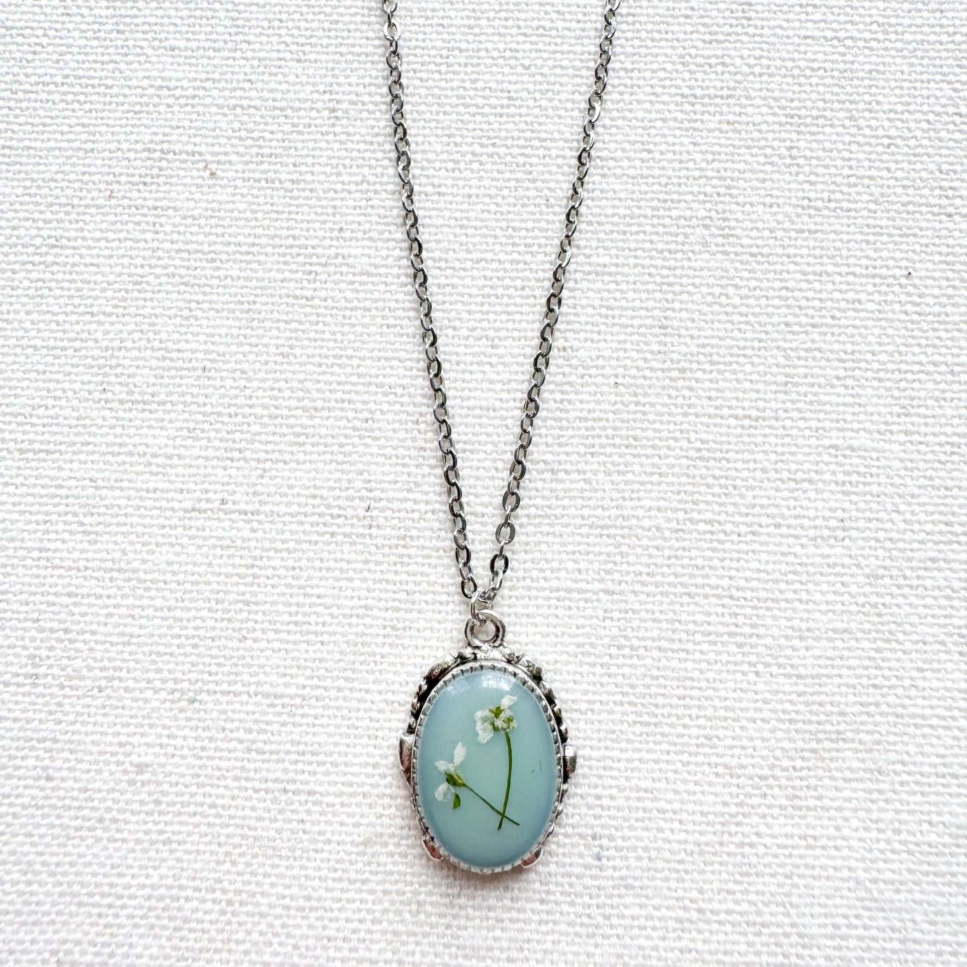 Handcrafted pressed flower pendant necklace featuring real dried Queen Anne's lace flowers against aquamarine backdrop, set in an ornate vintage silver frame. Perfect for nature-inspired jewelry lovers.