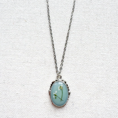 Handcrafted pressed flower pendant necklace featuring real dried Queen Anne's lace flowers against aquamarine backdrop, set in an ornate vintage silver frame. Perfect for nature-inspired jewelry lovers.