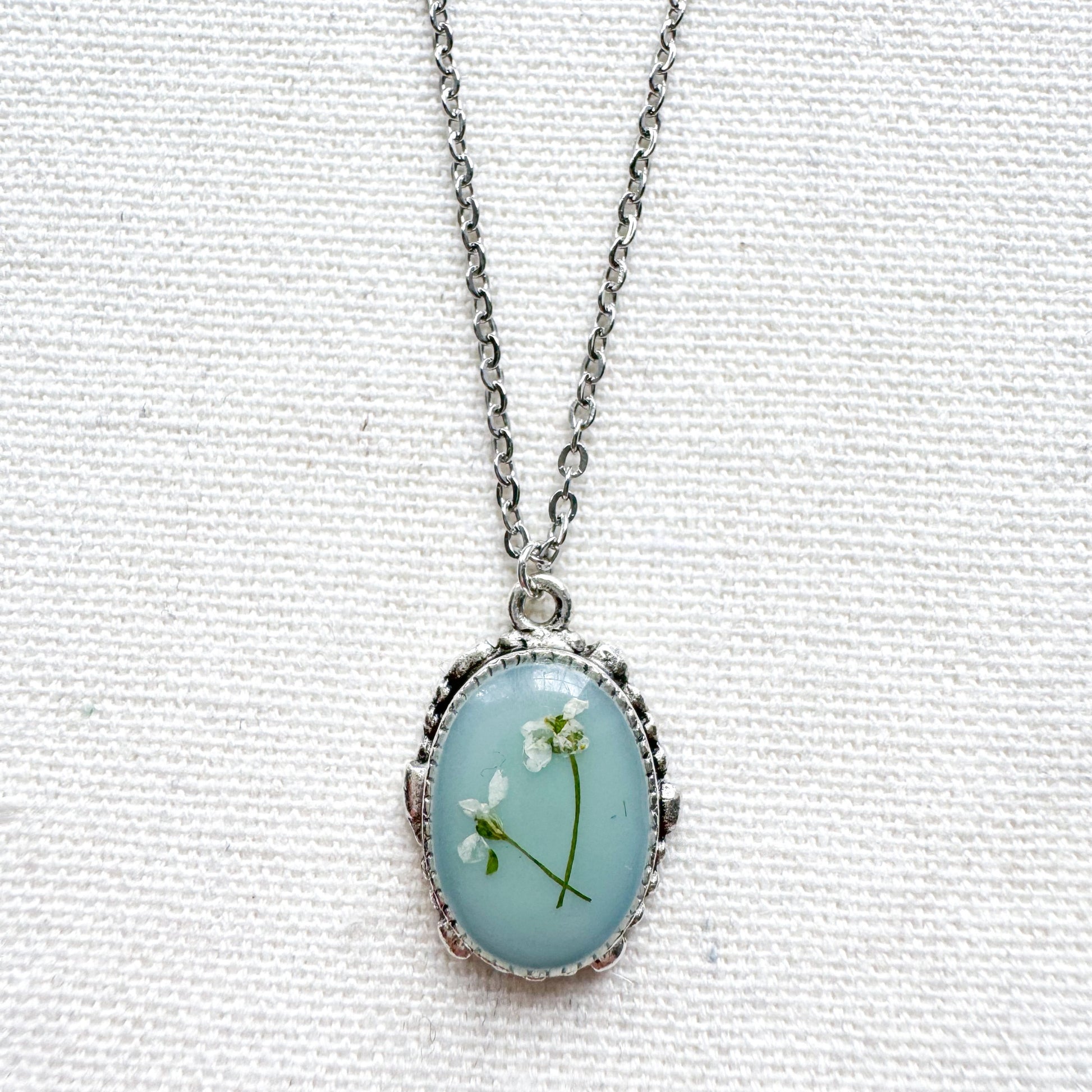 Handcrafted pressed flower pendant necklace featuring real dried Queen Anne's lace flowers against aquamarine backdrop, set in an ornate vintage silver frame. Perfect for nature-inspired jewelry lovers.