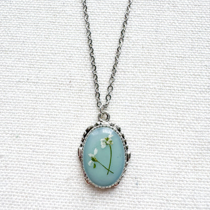 Handcrafted pressed flower pendant necklace featuring real dried Queen Anne's lace flowers against aquamarine backdrop, set in an ornate vintage silver frame. Perfect for nature-inspired jewelry lovers.