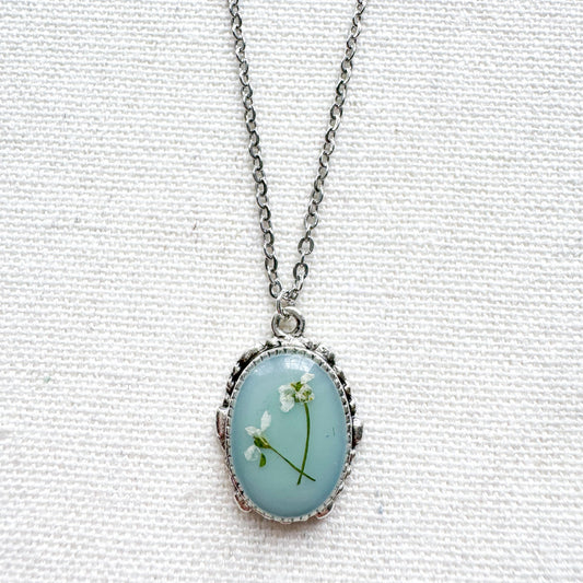 Handcrafted pressed flower pendant necklace featuring real dried Queen Anne's lace flowers against aquamarine backdrop, set in an ornate vintage silver frame. Perfect for nature-inspired jewelry lovers.