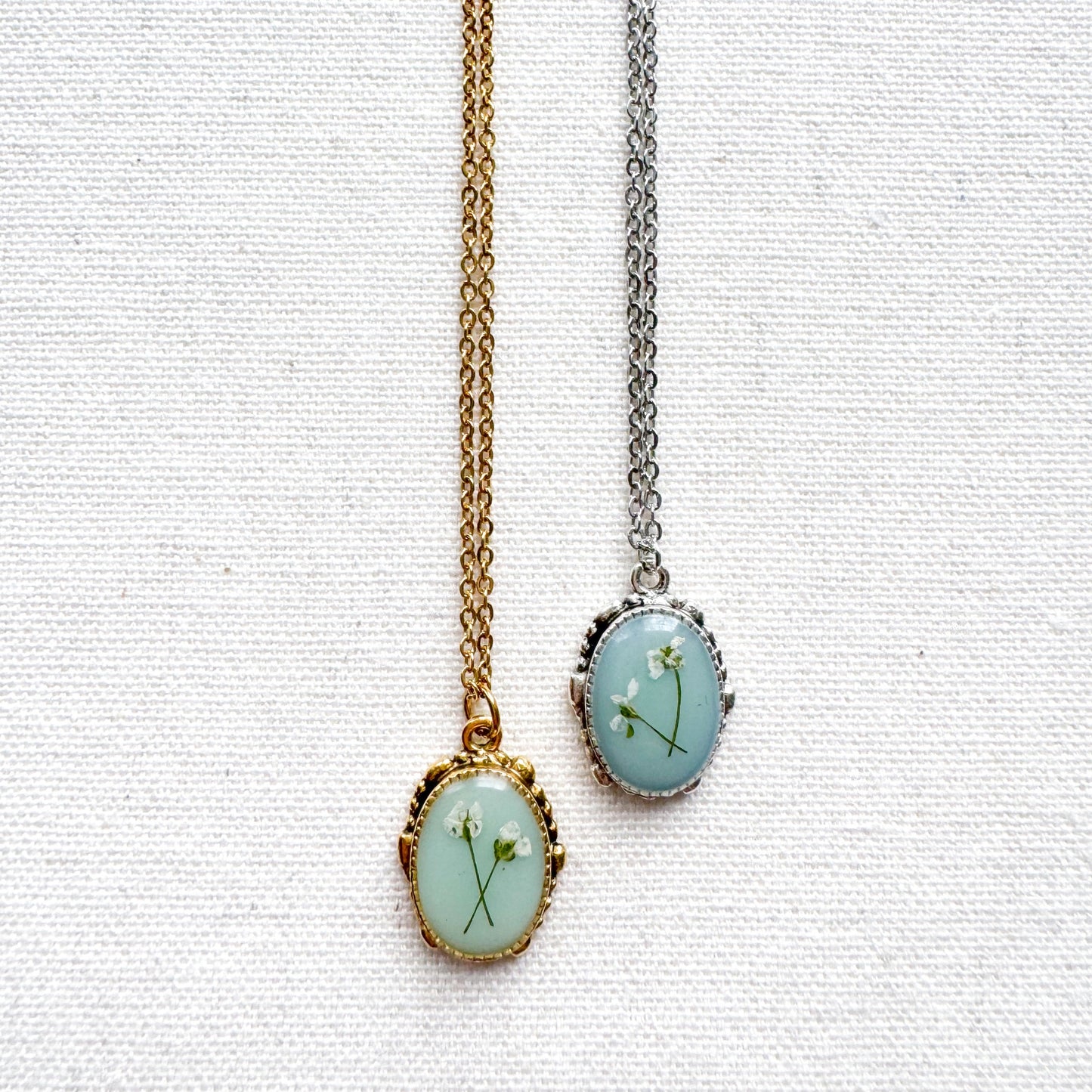 Two handcrafted pressed flower pendant necklaces featuring real dried Queen Anne's lace flowers against aquamarine backdrop. One is set in an ornate vintage silver pendant and the other antique gold pendant frame. Perfect for nature-inspired jewelry lovers.