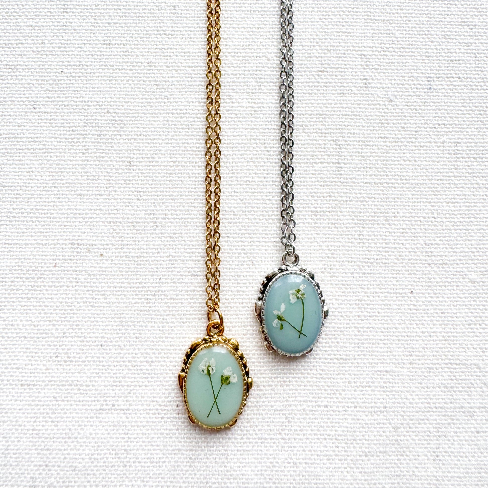 Two handcrafted pressed flower pendant necklaces featuring real dried Queen Anne's lace flowers against aquamarine backdrop. One is set in an ornate vintage silver pendant and the other antique gold pendant frame. Perfect for nature-inspired jewelry lovers.