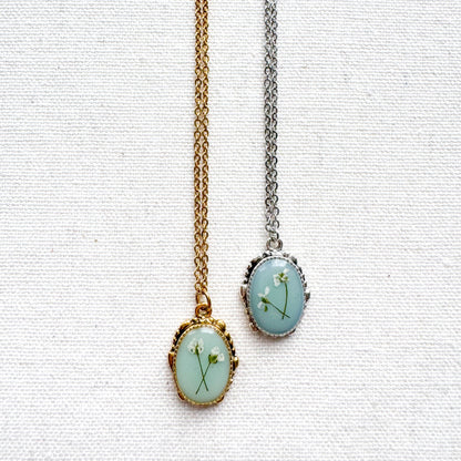 Two handcrafted pressed flower pendant necklaces featuring real dried Queen Anne's lace flowers against aquamarine backdrop. One is set in an ornate vintage silver pendant and the other antique gold pendant frame. Perfect for nature-inspired jewelry lovers.