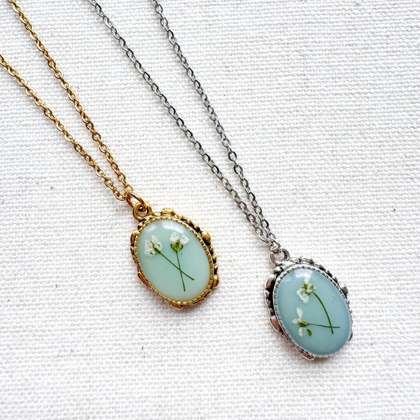 Two handcrafted pressed flower pendant necklaces featuring real dried Queen Anne's lace flowers against aquamarine backdrop. One is set in an ornate vintage silver pendant and the other antique gold pendant frame. Perfect for nature-inspired jewelry lovers.
