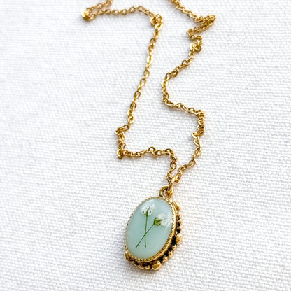 Handcrafted pressed flower pendant necklace featuring real dried Queen Anne's lace flowers against aquamarine backdrop, set in an ornate vintage gold frame, perfect for nature-inspired jewelry lovers.